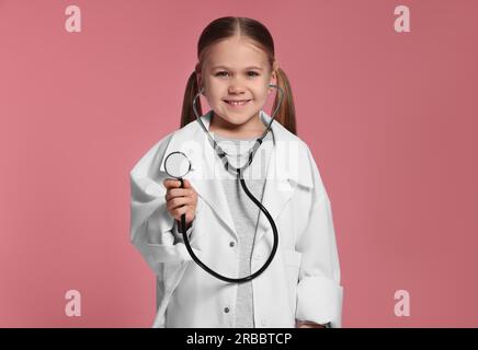 Bambina in uniforme medica con stetoscopio su sfondo rosa Foto Stock