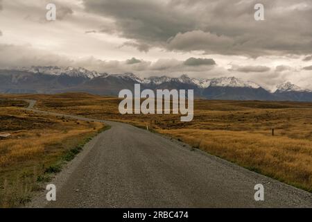 Viaggia verso le alpi meridionali innevate su una strada ghiaiosa e ventosa attraverso terreni agricoli rurali tra il lago Takapo e il lago Pukaki Foto Stock