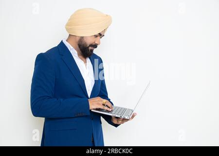 Giovane uomo d'affari indiano sikh con vestito blu e turbante che tiene il computer portatile isolato su sfondo bianco. Concetto aziendale. Foto Stock