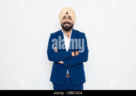 Felice giovane uomo d'affari indiano sikh che indossa una tuta in piedi con le braccia a croce isolato su sfondo bianco. Concetto aziendale. Foto Stock