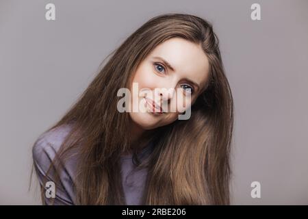 Modello dagli occhi blu, capelli lunghi scuri, espressione soddisfatta. Posa su sfondo grigio, sguardo diretto. Bellezza e persone. Foto Stock