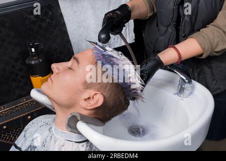 Le mani di una ragazza, un esperto lavoratore di salone con guanti neri, lavano via la vernice dai capelli del cliente di un giovane ragazzo con acqua e pioggia sopra Foto Stock