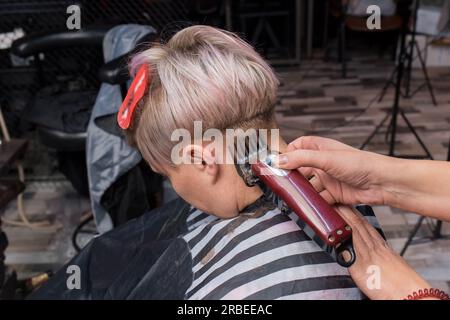 Le mani di una ragazza, esperta maestra di parrucchiere, nella sua attività professionale, tagliano i capelli di una donna cliente al lavoro con una macchina. Foto Stock