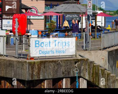 Persone che camminano nell'area dei moli dopo il cartello "gite in barca partono qui", Oban, Argyll and Bute, Scozia, Regno Unito. Foto Stock