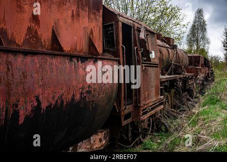 Locomotiva a steem arrugginita con carrozza a carbone abbandonata al cimitero dei treni sul vecchio binario ferroviario. Foto Stock