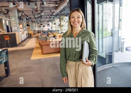 Bella direttrice con abiti casual in piedi con un computer portatile in un accogliente coworking e guardando la macchina fotografica Foto Stock