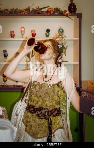 06-02-2019 Tulsa USA - donna vestita come ninfa boschiva o fata con fiori nei capelli che si abbassano un drink da un bicchiere di vino di legno in verde e crema Foto Stock