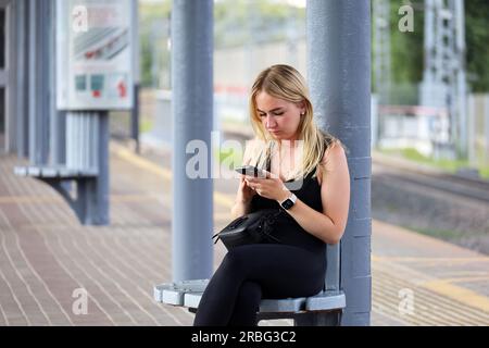 Ragazza bionda che usa uno smartphone seduto su una panchina della stazione ferroviaria in estate Foto Stock