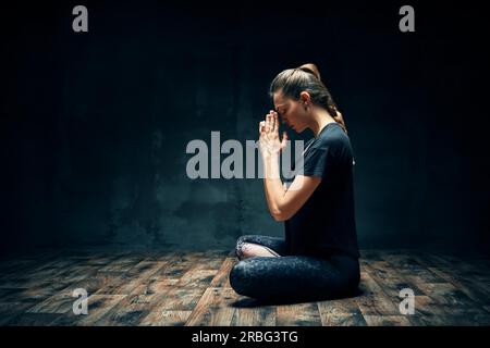 Vista posteriore di una giovane donna che pratica yoga seduta in posa di loto con un nome nella stanza buia e nello spazio per le copie. Esercizio Ardha Padmasana, concetto di meditazione Foto Stock