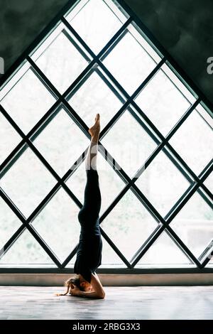 Giovane donna yogi attraente che pratica yoga in piedi nella posa sostenuta del poggiatesta, Salamba Sirsasana, su un grande sfondo triangolare della finestra. in salute Foto Stock