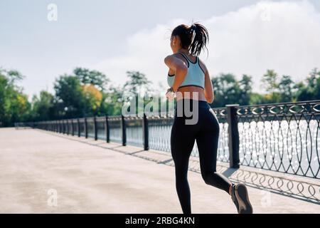 Vista posteriore di una donna in forma che corre all'aperto lungo il fiume. Bella donna con abbigliamento sportivo che fa jogging all'aperto. Movimento dinamico Foto Stock