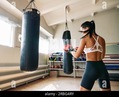 Vista posteriore del pugile femminile che colpisce un sacco di punzonatura enorme in palestra fitness. Donna che pratica i suoi pugni in uno studio di boxe Foto Stock