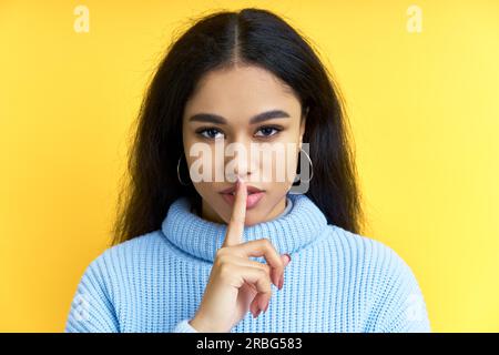 Bella donna nera tiene il dito sulle labbra, facendo gesto hush su sfondo giallo. Shh, concetto di silenzio Foto Stock