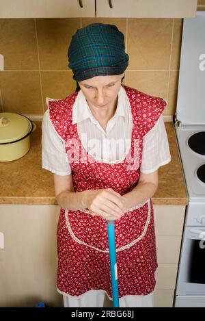 Una donna adulta depressa, una casalinga o una cameriera, che indossa un grembiule rosso e una sciarpa verde sulla testa, riposa dopo aver spazzato la cucina con un Foto Stock