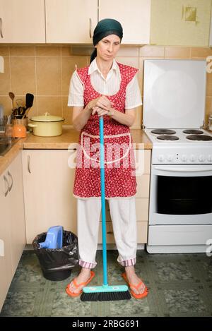 Una donna adulta depressa, una casalinga o una cameriera, che indossa un grembiule rosso e una sciarpa verde sulla testa, riposa dopo aver spazzato la cucina con un Foto Stock