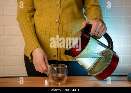 Donna che usa l'acqua bollita bollita dal bollitore elettrico in una tazza di vetro trasparente sul tavolo. Processo di preparazione del tè o cerimonia del tè in una luce calda e soffusa. M Foto Stock