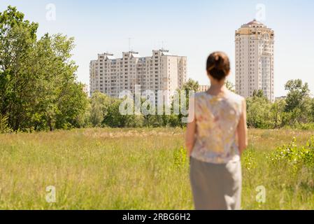 Donna sfocata che guarda moderni edifici residenziali mentre guarda le sue case in campagna, in mezzo ai prati, sotto il sole durante un caldo Foto Stock