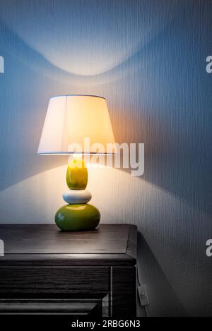 Piccola lampada gialla, grigia e verde su un comò in legno o su un tavolo da notte, che illumina la parete coperta di carta da parati di notte o di sera tardi. Morbido Foto Stock