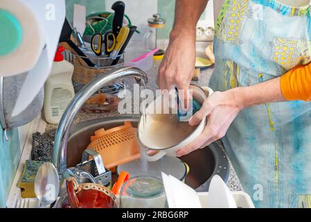 Un uomo sta lavando i piatti in cucina. Sta usando una spugna e un prodotto per lavare i piatti. L'acqua calda scorre dal rubinetto Foto Stock