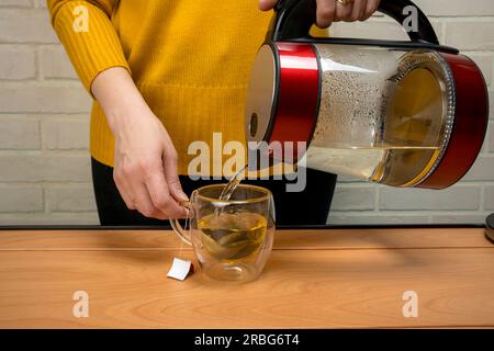 Donna che usa l'acqua bollita bollita dal bollitore elettrico in una tazza di vetro trasparente sul tavolo. Processo di preparazione del tè o cerimonia del tè in una luce calda e soffusa. M Foto Stock