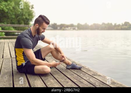 Attraente barbuto giovane uomo relax su un ponte di legno che si affaccia su di un lago calmo con il suo telefono cellulare in mano guardando la telecamera con un sorriso Foto Stock