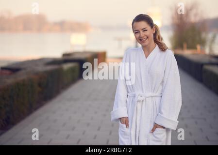 Giovane donna sorridente in un accappatoio bianco in piedi all'aperto su una passerella pavimentata che conduce a un lago tranquillo con le mani nelle tasche per un benessere Foto Stock