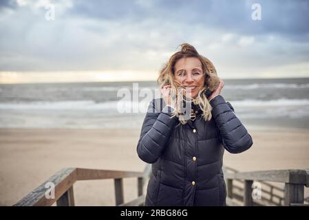 Donna di mezza età che affronta una fredda giornata invernale al mare in piedi su una terrazza di legno che si affaccia sulla spiaggia in una giornata ventilata sorridente Foto Stock