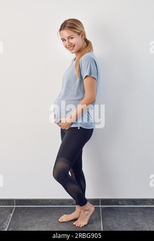 Vista laterale di un felice casual a piedi nudi incinta donna giovane cullano il suo bambino urtare con le sue mani svolta a guardare la fotocamera con un caldo sorriso Foto Stock