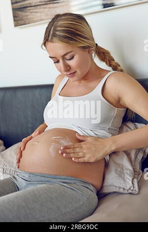 Giovane donna incinta sfregamento crema idratante sul suo ventre per idratare la sua pelle e ridurre la possibilità di smagliature dopo il parto Foto Stock