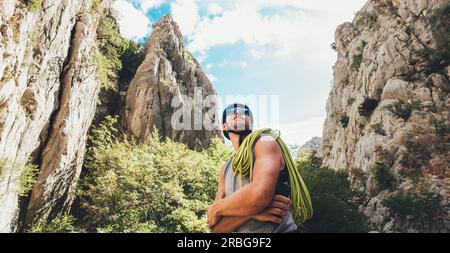 Ritratto di sorridente arrampicatore in casco protettivo e occhiali da sole con corda da arrampicata sulla spalla in piedi nel parco Paklenica tra roccia w rupe Foto Stock