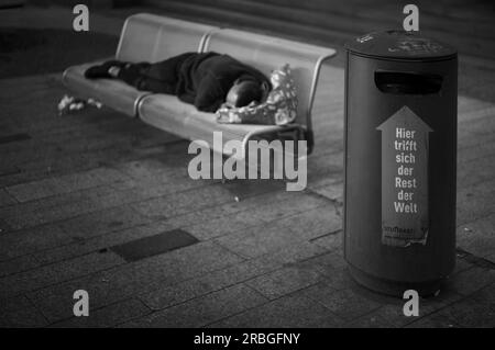 Un senzatetto, berbero, sdraiato sulla panchina del parco, dormendo, di fronte ad esso, bidone della spazzatura con scritta QUI INCONTRA IL RESTO DEL MONDO, Stoccarda Foto Stock