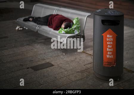 Un senzatetto, berbero, sdraiato sulla panchina del parco, dormendo, di fronte ad esso, bidone della spazzatura con scritta QUI INCONTRA IL RESTO DEL MONDO, Stoccarda Foto Stock