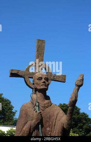 Parte della statua di Sant'Aidan nel Priorato Lindisfarne dell'artista Kathleen Parbury. Statua rossa in cemento di St Aidan con torcia, truffatore di bronzo, croce. Foto Stock