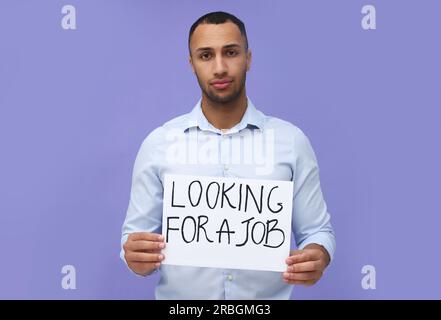 Giovane disoccupato con un cartello con una frase che cerca Un lavoro su sfondo violetto Foto Stock