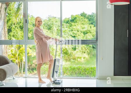 La giovane donna incinta ama pulire la sua casa. Facile pulizia con un aspirapolvere wireless. Donna incinta che pulisce il pavimento con l'aspirapolvere portatile Foto Stock