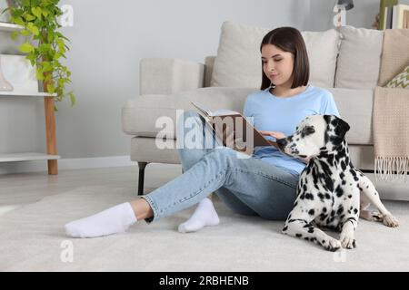 Bella giovane donna che legge un libro e il suo adorabile cane dalmata sul pavimento a casa. Adorabile animale domestico Foto Stock