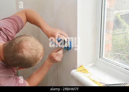 In primo piano, l'elettricista sta installando una nuova presa elettrica, una presa multi-spina, alimentando i fili attraverso le scatole elettriche in plastica verso una nuova presa. Foto Stock