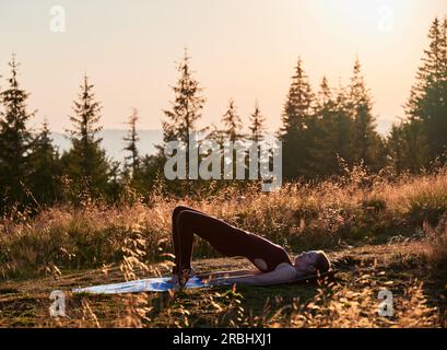 Bella donna che fa una posa a ponte a una gamba. Allenati all'aperto la mattina presto. Esercizio all'aria aperta. Foto Stock