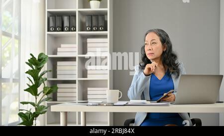 Una donna d'affari asiatica premurosa e seria o un capo donna che guarda fuori dalla finestra con un volto serio, pianificando la sua attività mentre si siede seduti Foto Stock