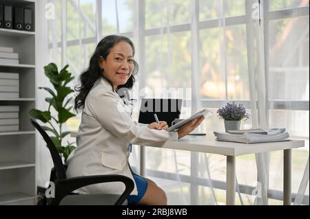 Un ritratto di una bella e sorridente donna d'affari asiatica matura o di un capo donna in un abito formale, che guarda la macchina fotografica, seduto alla sua scrivania in lei Foto Stock