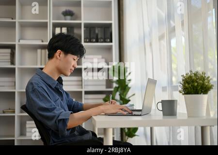 Un giovane lavoratore d'ufficio asiatico serio e concentrato sta lavorando sul suo computer portatile alla sua scrivania in un ufficio moderno. Foto Stock