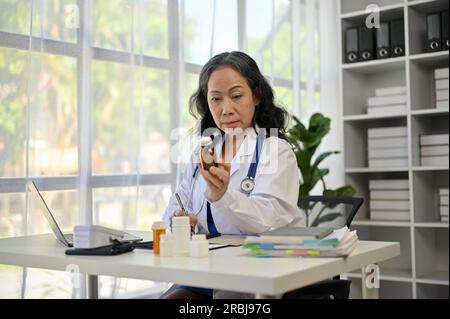 Una professionista asiatica anziana medico si concentra sulla scrittura di prescrizioni mediche e lavorare alla sua scrivania nello studio dell'ospedale. Operatore medico e. Foto Stock