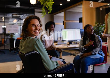 Ritratto di donne d'affari caucasiche felici in sedia a rotelle che tengono un tablet in ufficio. Ufficio informale, lavoro di squadra, comunicazione, disabilità, Foto Stock