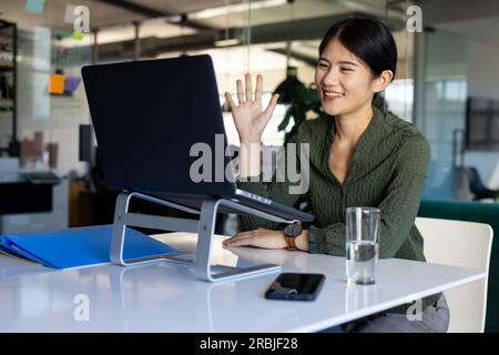 Felice donna d'affari asiatica alla scrivania in ufficio che videochiama sul computer portatile, salutando. Lavoro, affari, comunicazione e ufficio informale, inalterato. Foto Stock