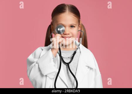 Bambina in uniforme medica con stetoscopio su sfondo rosa Foto Stock