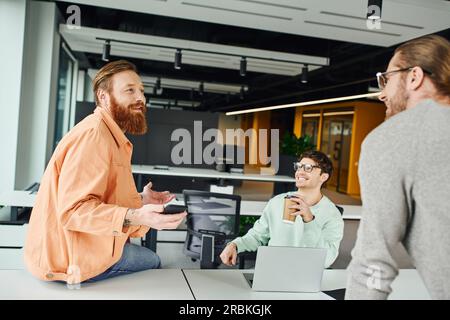 uomo barbuto ispirato che tiene uno smartphone e discute di un nuovo progetto aziendale con i colleghi vicino a un computer portatile sulla scrivania di lavoro in un moderno ufficio di coworking, Foto Stock