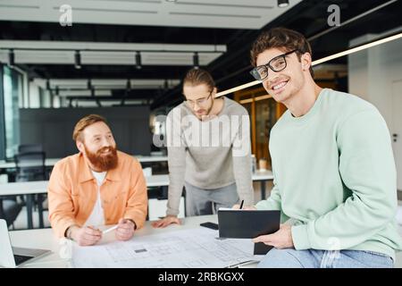 uomo d'affari soddisfatto con occhiali che tiene un notebook e guarda la fotocamera accanto a architetti creativi che lavorano con blueprint vicino a un notebook in contemporanea Foto Stock