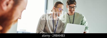 architetti creativi in abiti casual e occhiali da vista che guardano un computer portatile vicino a un collega che lavora su foreground sfocato, collaborazione, idee innovative, Foto Stock