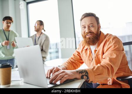 allegro architetto barbuto e tatuato che lavora su un computer portatile vicino al punto da asporto beve in un bicchiere di carta e guarda la fotocamera mentre i colleghi parlano di b sfocata Foto Stock