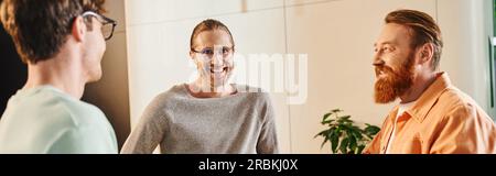 uomo d'affari con la barba gioiosa sorridente vicino a colleghi ispirati in occhiali che parlano durante la riunione nella sala dello spazio di coworking contemporaneo, partnership Foto Stock